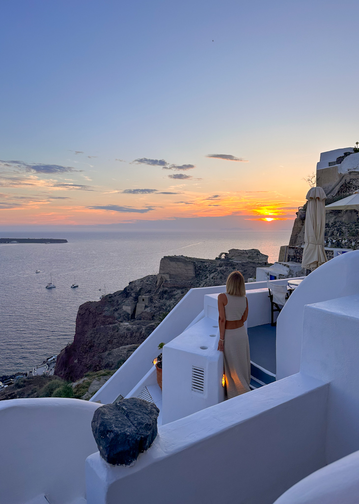 Dove alloggiare a Santorini - ragazza su una terrazza con mare e tramonto a Santorini